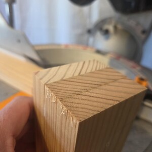 May include: Close-up of a light brown wooden block with angled cuts, likely for woodworking. The block has visible wood grain and is held near a saw, suggesting a DIY or construction project.