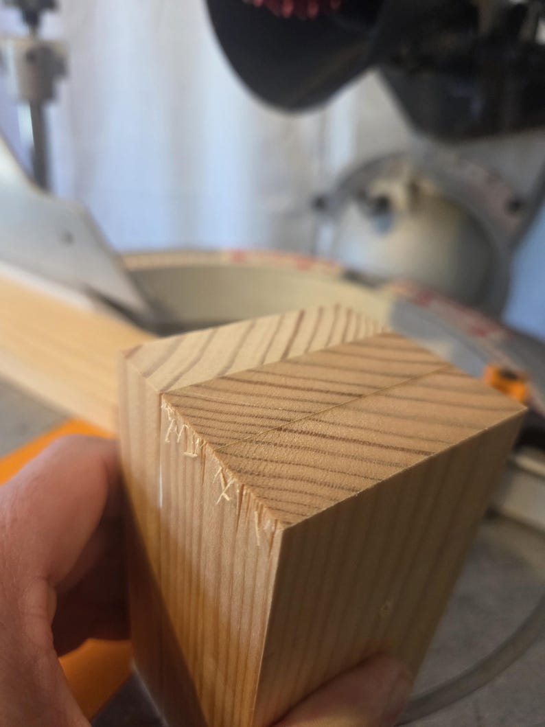 May include: Close-up of a light brown wooden block with angled cuts, likely for woodworking. The block has visible wood grain and is held near a saw, suggesting a DIY or construction project.