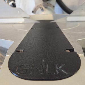 May include: Close-up of a black, textured surface with the letters "GNLK" embossed on it. The surface is part of a silver and gray power tool, likely a miter saw, with a blade and other components visible in the background.