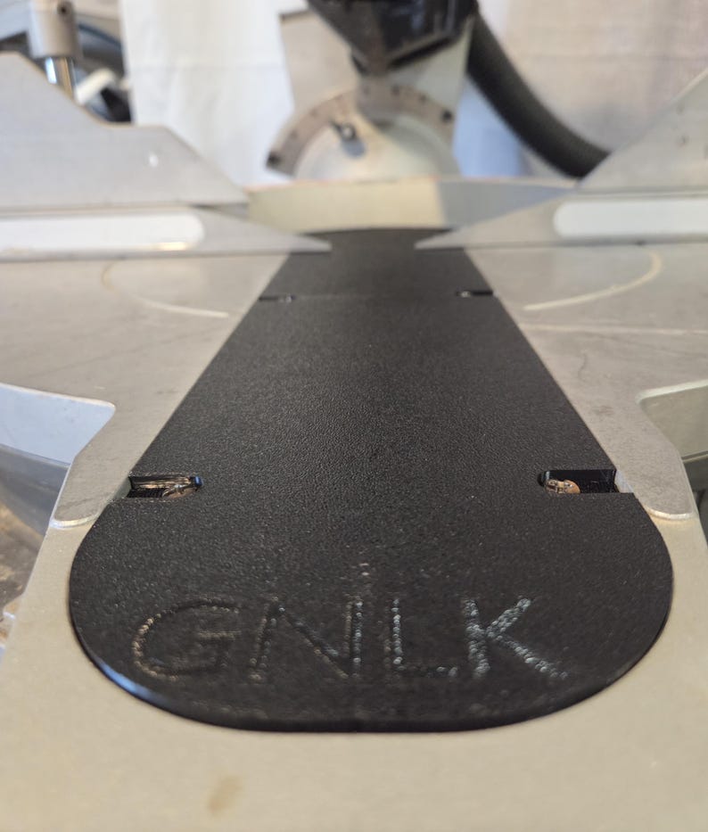 May include: Close-up of a black, textured surface with the letters "GNLK" embossed on it. The surface is part of a silver and gray power tool, likely a miter saw, with a blade and other components visible in the background.