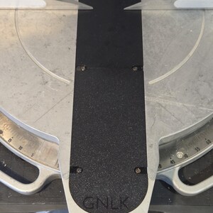 May include: A close-up view of a miter saw with a black textured surface and silver metal components. The saw features a degree scale and the letters "GNLK" are visible on the base.