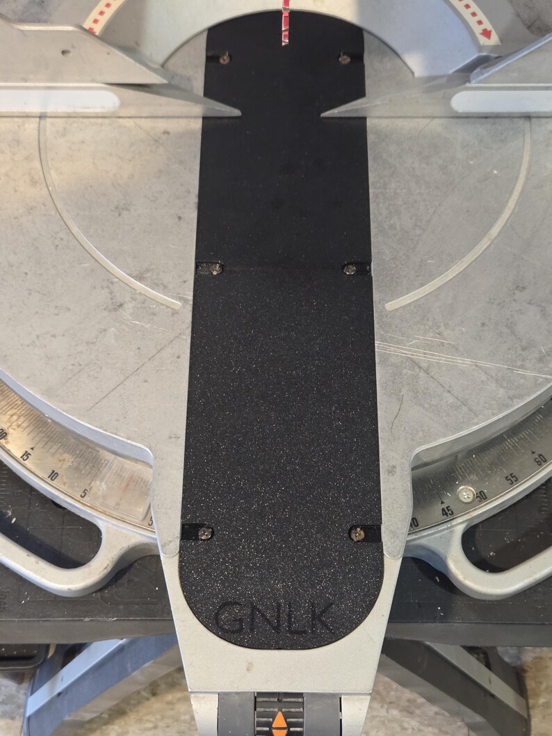 May include: A close-up view of a miter saw with a black textured surface and silver metal components. The saw features a degree scale and the letters "GNLK" are visible on the base.