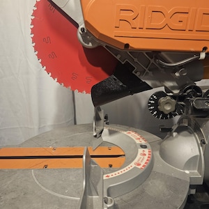 K&ouml;nnte beinhalten: Eine orangefarbene und silberne Gehrungss&auml;ge mit rotem S&auml;geblatt. Auf der S&auml;ge ist der Markenname "RIDGID" aufgedruckt. Das kreisf&ouml;rmige S&auml;geblatt hat scharfe Z&auml;hne. Dieses Werkzeug ist zum Schneiden von Holz und anderen Materialien bestimmt.