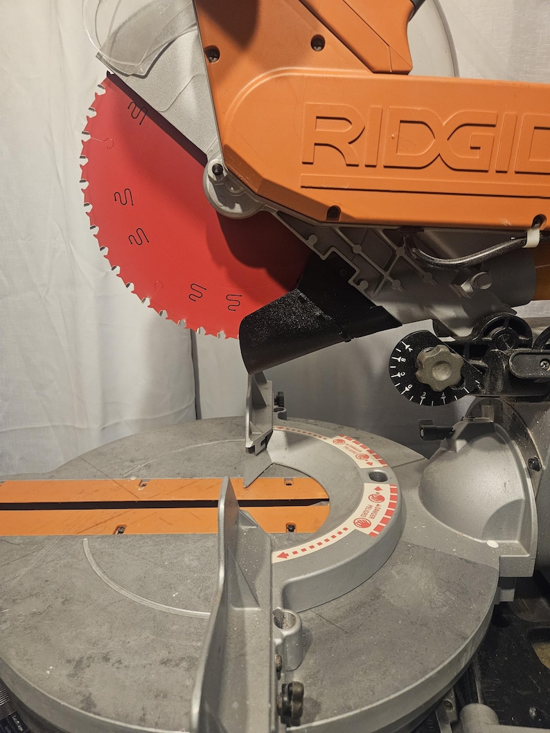 K&ouml;nnte beinhalten: Eine orangefarbene und silberne Gehrungss&auml;ge mit rotem S&auml;geblatt. Auf der S&auml;ge ist der Markenname "RIDGID" aufgedruckt. Das kreisf&ouml;rmige S&auml;geblatt hat scharfe Z&auml;hne. Dieses Werkzeug ist zum Schneiden von Holz und anderen Materialien bestimmt.