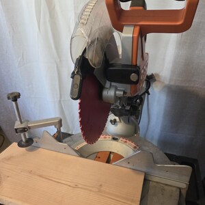 May include: An orange and silver miter saw with a red blade is shown with a piece of light brown wood. The saw has a clear plastic blade guard and a black handle. The wood is positioned on the saw's base, ready for cutting.