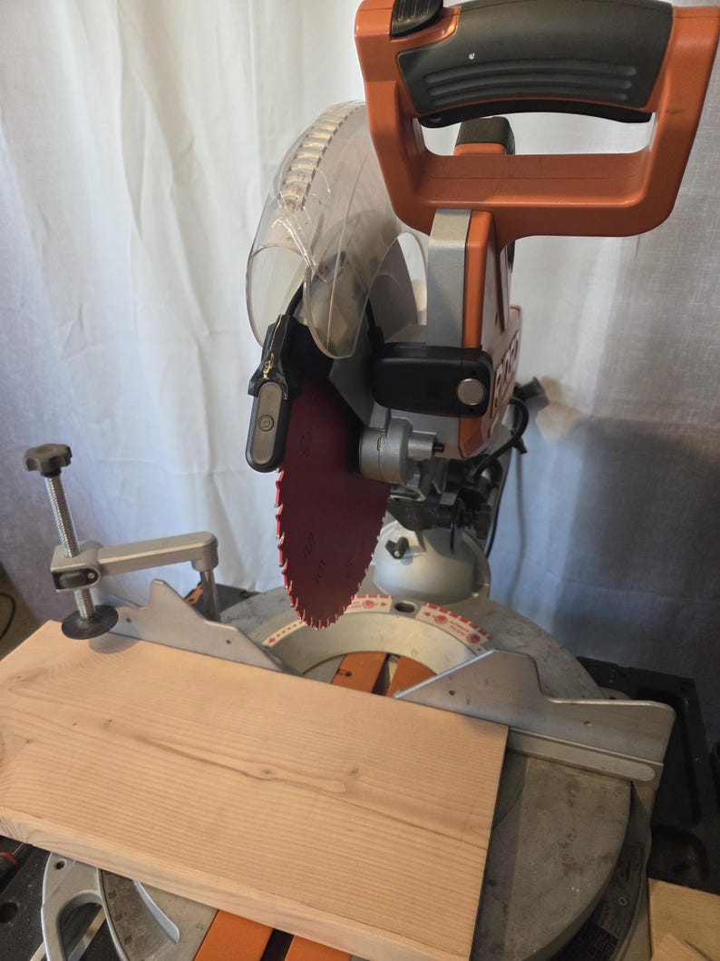 May include: An orange and silver miter saw with a red blade is shown with a piece of light brown wood. The saw has a clear plastic blade guard and a black handle. The wood is positioned on the saw's base, ready for cutting.