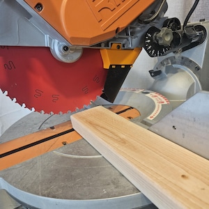 K&ouml;nnte beinhalten: Nahaufnahme einer Gehrungss&auml;ge mit orangefarbenem Geh&auml;use und rotem S&auml;geblatt. Ein St&uuml;ck helles Holz liegt auf der Basis der S&auml;ge und ist bereit zum Schneiden. Die S&auml;ge hat eine silberne Basis und ein schwarzes Netzkabel.