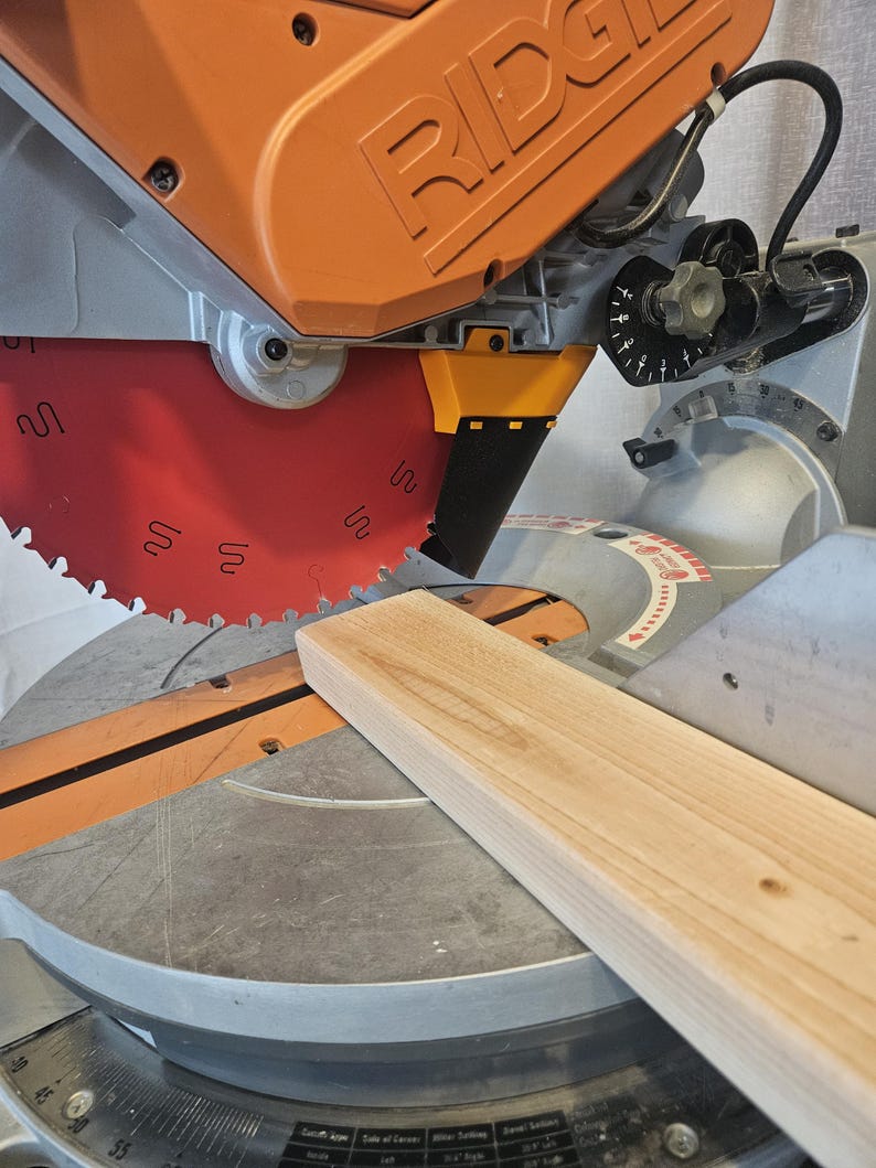K&ouml;nnte beinhalten: Nahaufnahme einer Gehrungss&auml;ge mit orangefarbenem Geh&auml;use und rotem S&auml;geblatt. Ein St&uuml;ck helles Holz liegt auf der Basis der S&auml;ge und ist bereit zum Schneiden. Die S&auml;ge hat eine silberne Basis und ein schwarzes Netzkabel.