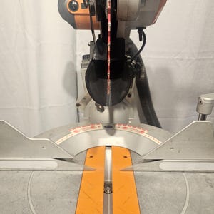K&ouml;nnte beinhalten: Eine silber-orangefarbene Gehrungss&auml;ge mit rotem S&auml;geblatt. Die S&auml;ge hat einen kreisf&ouml;rmigen Klingenschutz und eine Basis mit einer orangefarbenen F&uuml;hrung. Die S&auml;ge ist zum Schneiden von Holz und anderen Materialien bestimmt.