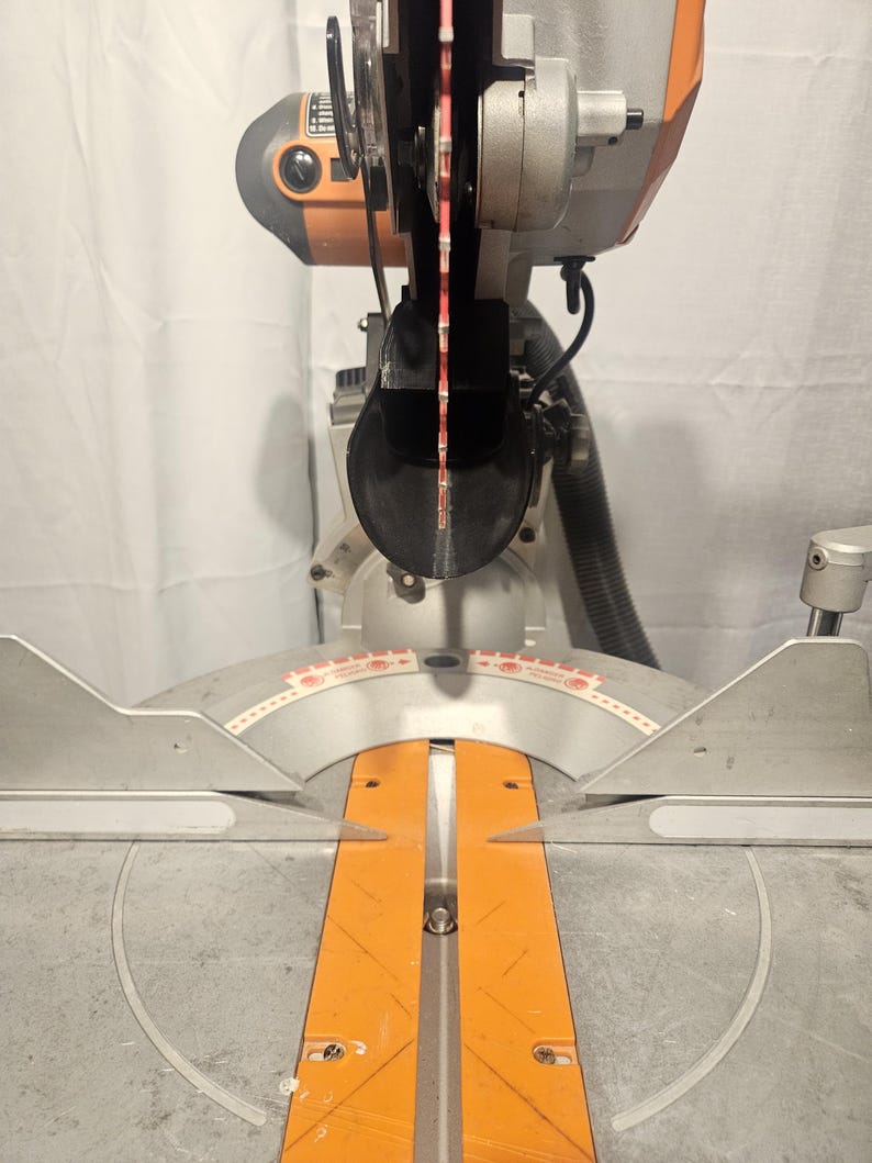 K&ouml;nnte beinhalten: Eine silber-orangefarbene Gehrungss&auml;ge mit rotem S&auml;geblatt. Die S&auml;ge hat einen kreisf&ouml;rmigen Klingenschutz und eine Basis mit einer orangefarbenen F&uuml;hrung. Die S&auml;ge ist zum Schneiden von Holz und anderen Materialien bestimmt.