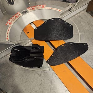 K&ouml;nnte beinhalten: Schwarze Gehrungss&auml;gen-Teile, darunter zwei strukturierte Klingenschutzvorrichtungen und ein schwarzes Kunststoffteil, sind auf einer silbernen und orangefarbenen Gehrungss&auml;ge angeordnet. Das S&auml;geblatt ist im Hintergrund sichtbar.