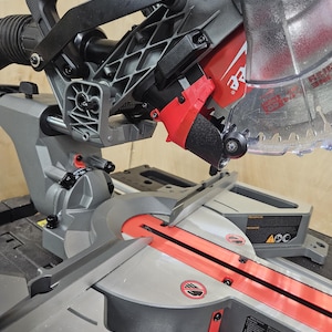 May include: A red and grey power mitre saw with a black handle and a clear plastic guard. The saw is on a table with a red and grey base. The saw has a warning label that says "Caution: Keep hands clear of blade".