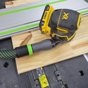 Puede incluir: Una lijadora DeWalt XR 20V Brushless amarilla y negra con una manguera verde y negra. La lijadora está sobre una superficie de madera.