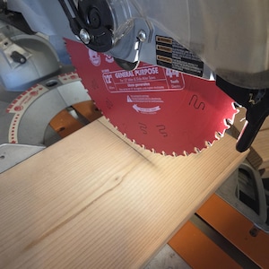 May include: A red 12-inch general purpose saw blade is positioned above a light-colored wooden plank. The saw is part of a miter saw, with a clear safety guard and black and silver components. The blade has 44 teeth.