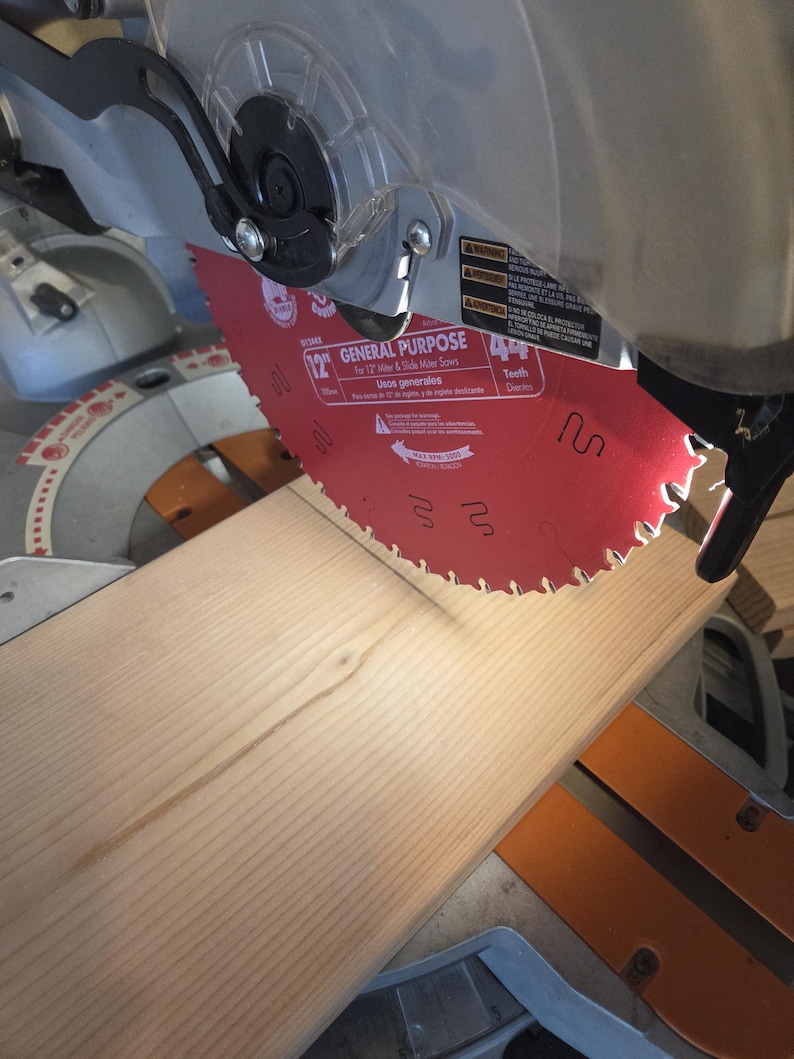 May include: A red 12-inch general purpose saw blade is positioned above a light-colored wooden plank. The saw is part of a miter saw, with a clear safety guard and black and silver components. The blade has 44 teeth.
