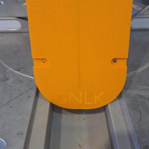 May include: Close-up of an orange, rectangular object with rounded edges. The object has two small holes near the top and the letters "GNLK" are embossed near the bottom. The object is set against a gray surface.