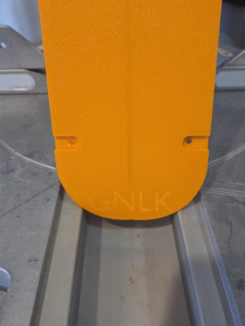 May include: Close-up of an orange, rectangular object with rounded edges. The object has two small holes near the top and the letters "GNLK" are embossed near the bottom. The object is set against a gray surface.
