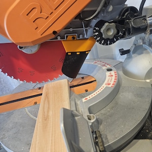 K&ouml;nnte beinhalten: Eine orangefarbene und graue Gehrungss&auml;ge mit einem roten S&auml;geblatt, das ein St&uuml;ck hellbraunes Holz schneidet. Die S&auml;ge hat ein schwarzes Zifferblatt und eine Schutzvorrichtung. Das Holz ist f&uuml;r einen pr&auml;zisen Schnitt positioniert.