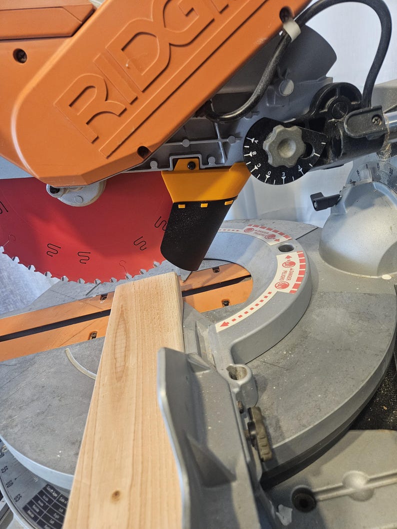 K&ouml;nnte beinhalten: Eine orangefarbene und graue Gehrungss&auml;ge mit einem roten S&auml;geblatt, das ein St&uuml;ck hellbraunes Holz schneidet. Die S&auml;ge hat ein schwarzes Zifferblatt und eine Schutzvorrichtung. Das Holz ist f&uuml;r einen pr&auml;zisen Schnitt positioniert.