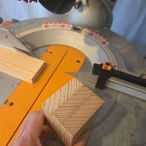 May include: A close-up of a wooden block with a mitered corner, held near a miter saw. The saw has an orange base and a red blade. A piece of wood is also on the saw's base.