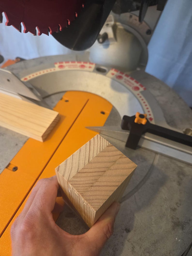 May include: A close-up of a wooden block with a mitered corner, held near a miter saw. The saw has an orange base and a red blade. A piece of wood is also on the saw's base.