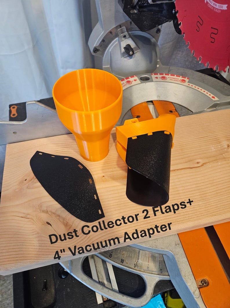 K&ouml;nnte beinhalten: Ein orangefarbener, 3D-gedruckter Staubabscheider mit zwei schwarzen Klappen und einem 10,16 cm Vakuumadapter. Der Text "Dust Collector 2 Flaps+ 4" Vacuum Adapter" ist auf die Holzoberfl&auml;che gedruckt. Ein rotes S&auml;geblatt ist sichtbar.