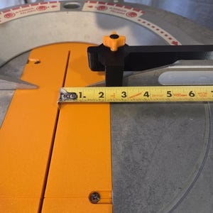 May include: A close-up of a miter saw with an orange and black design. The blade guard is clear. A yellow measuring tape shows a measurement of approximately 3 inches.