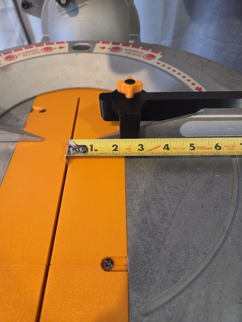 May include: A close-up of a miter saw with an orange and black design. The blade guard is clear. A yellow measuring tape shows a measurement of approximately 3 inches.