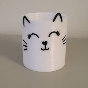 Op de afbeelding: Witte keramische potloodhouder in de vorm van een kat met zwarte getekende kenmerken, waaronder ogen, neus en snorharen. De kat glimlacht.