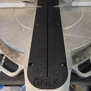 May include: A silver and black miter saw with a black base and a silver blade guard. The saw has a black center section with the letters "GNLK" at the bottom. The saw has a measurement scale.