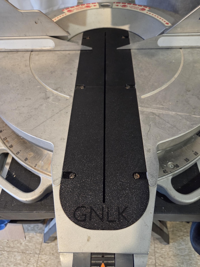May include: A silver and black miter saw with a black base and a silver blade guard. The saw has a black center section with the letters "GNLK" at the bottom. The saw has a measurement scale.