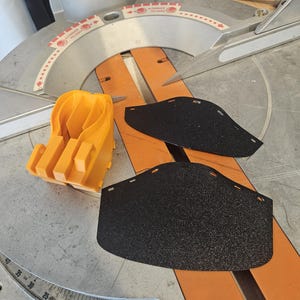 K&ouml;nnte beinhalten: Draufsicht auf eine Gehrungss&auml;ge mit einem orangefarbenen Kunststoffschutz und zwei schwarzen Schleifpads. Die S&auml;ge hat ein silbernes S&auml;geblatt und eine Messskala. Der orangefarbene Schutz befindet sich links.