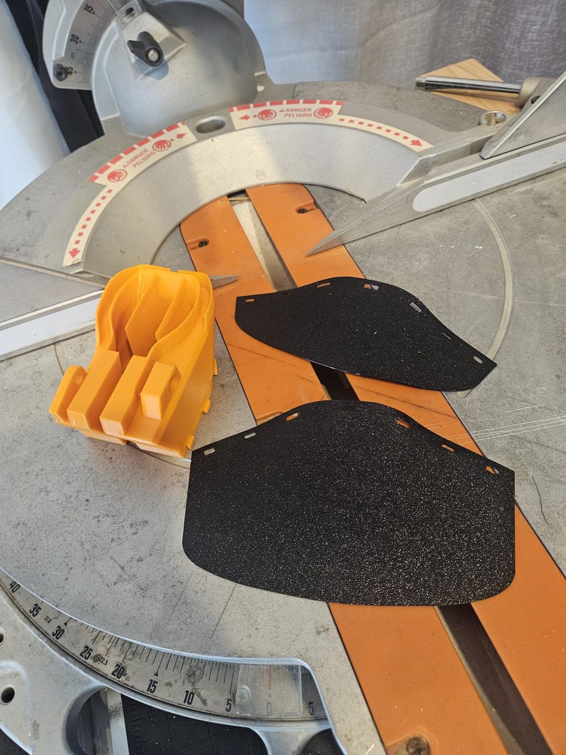 K&ouml;nnte beinhalten: Draufsicht auf eine Gehrungss&auml;ge mit einem orangefarbenen Kunststoffschutz und zwei schwarzen Schleifpads. Die S&auml;ge hat ein silbernes S&auml;geblatt und eine Messskala. Der orangefarbene Schutz befindet sich links.