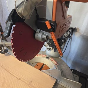 May include: A Ridgid miter saw with a red blade is positioned over a light brown wooden plank. An orange marker rests on the saw. The saw has a clear safety guard and an orange handle. The saw is designed for cutting wood.