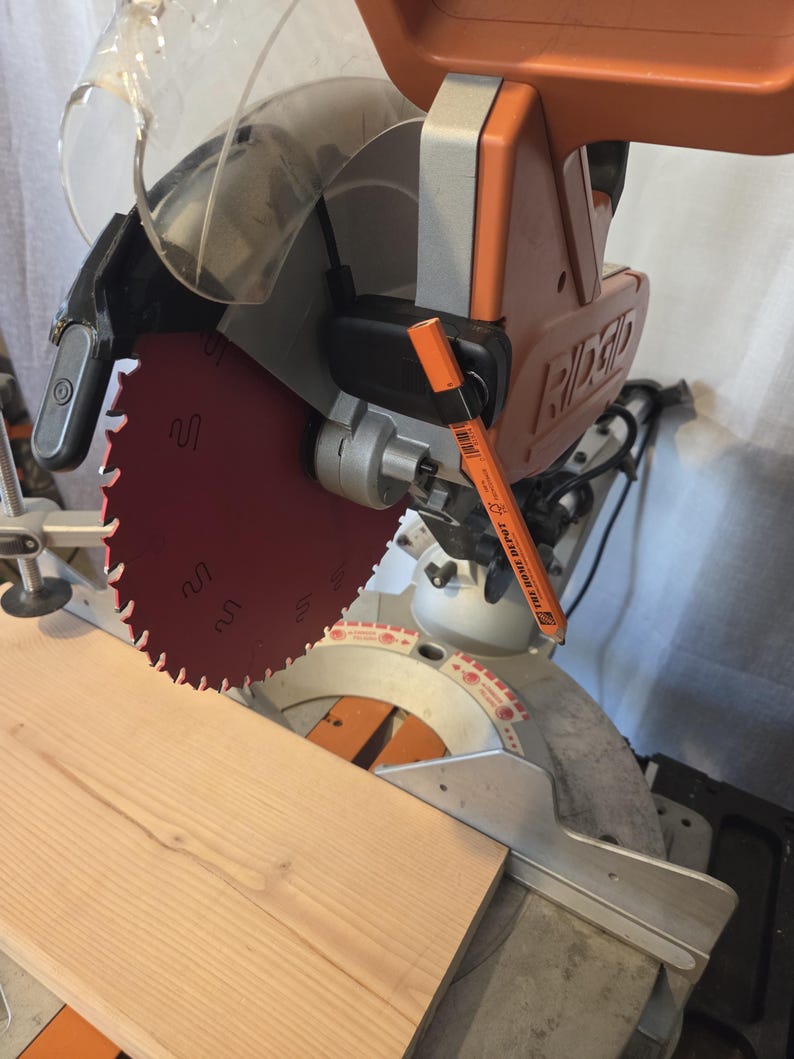 May include: A Ridgid miter saw with a red blade is positioned over a light brown wooden plank. An orange marker rests on the saw. The saw has a clear safety guard and an orange handle. The saw is designed for cutting wood.