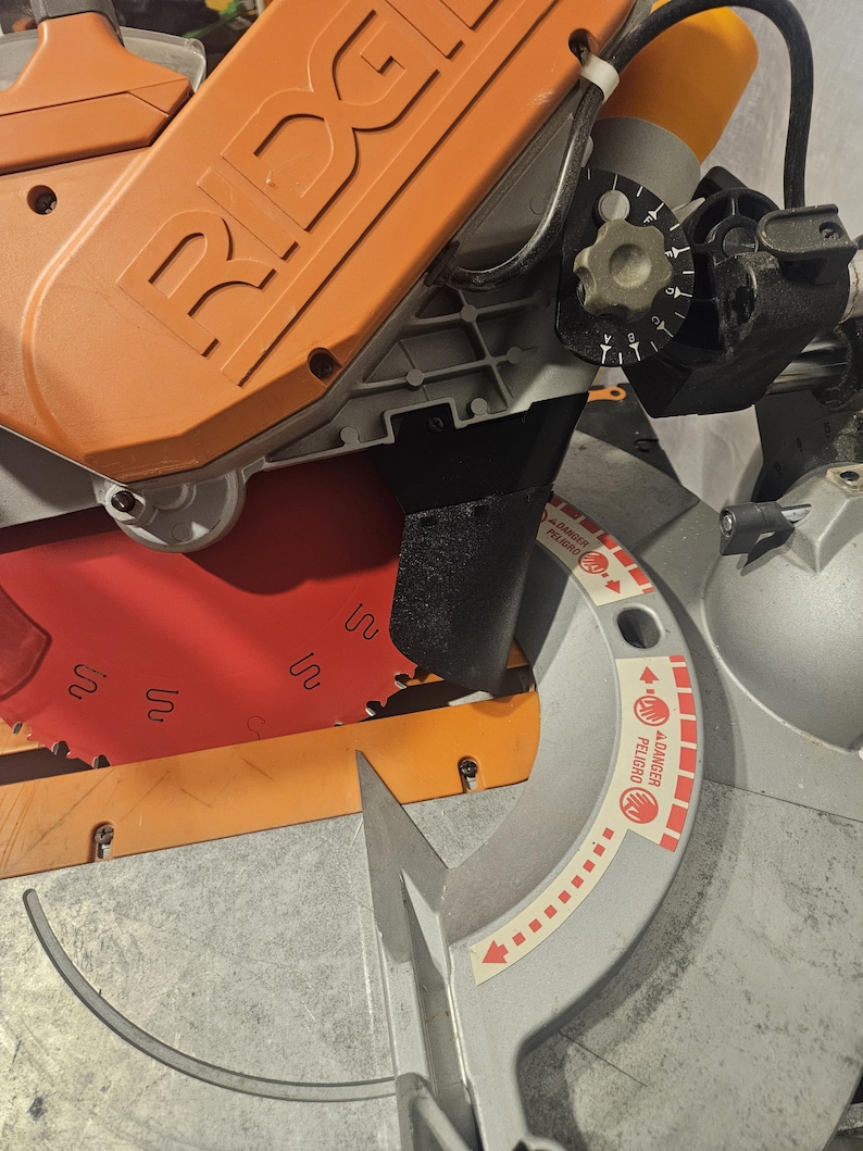 K&ouml;nnte beinhalten: Eine orangefarbene und graue Gehrungss&auml;ge mit rotem S&auml;geblatt. Auf dem orangefarbenen Geh&auml;use ist der Markenname "RIDGID" eingepr&auml;gt. Der Klingenschutz tr&auml;gt einen Warnhinweis "DANGER PELIGRO".