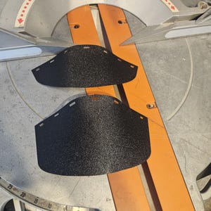 K&ouml;nnte beinhalten: Zwei schwarze, strukturierte Schutzvorrichtungen liegen auf einer orangefarbenen und silbernen Gehrungss&auml;ge. Die Schutzvorrichtungen haben eine gebogene Form mit einer Reihe kleiner L&ouml;cher am oberen Rand. Die S&auml;ge hat einen Klingenschutz und eine Messskala.