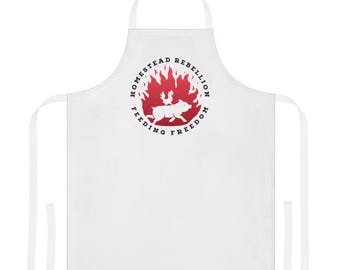 May include: White apron featuring a red circular design with the words "HOMESTEAD REBELLION" and "FEEDING FREEDOM". The design includes a pig, a chicken, and flames. The apron has a neck loop and waist ties.
