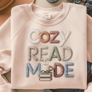 May include: A light pink sweatshirt with the words "COZY READ MADE" embroidered in various colors. The design includes a coffee cup, glasses, and books, suggesting a reading theme. The sweatshirt is a comfortable, casual style.