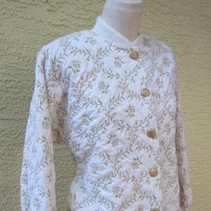 May include: A white long-sleeved jacket with a floral pattern and gold buttons. The jacket has a stand-up collar and a ribbed hem.