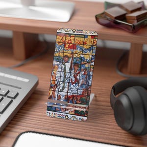 May include: A phone stand featuring a stained-glass design with figures in white robes, set against a backdrop of red, yellow, and blue. The stand is rectangular with a patterned border and is displayed on a wooden desk.