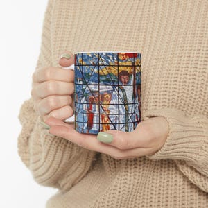 May include: White ceramic mug with a colorful stained glass design featuring a group of people. The design is reminiscent of a religious scene.