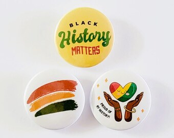 May include: Three round buttons with different designs. The first button is yellow with the text "Black History Matters" in green and black lettering. The second button is white with a multi-colored stripe design. The third button is white with a heart-shaped design in the colors of the Pan-African flag and the text "Proud of My History" in black lettering.