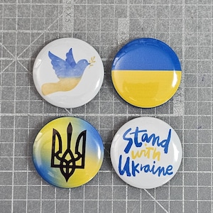 May include: Four circular pin badges with Ukrainian designs. One shows a blue dove with a yellow olive branch. Another has the Ukrainian flag. A third has a yellow and blue emblem. The last says "Stand with Ukraine."
