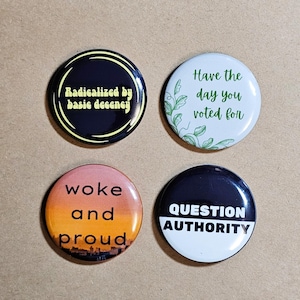 Może przedstawiać: Cztery okrągłe guziki z tekstem. Jeden mówi "Radicalized by basic decency" w kolorze żółtym na czarnym tle. Inny mówi "Have the day you voted for" z zielonymi liśćmi. Trzeci mówi "woke and proud" z projektem zachodu słońca. Ostatni mówi "QUESTION AUTHORITY".