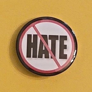 May include: A round button with a white background and the word "HATE" in bold black letters, crossed out with a red diagonal line. The button has a black border and is set against a yellow background.