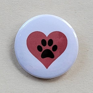 May include: A round, white button with a red heart and a black paw print in the center. The button is a simple design, perfect for pet lovers or anyone who loves animals. The button is likely made of metal or plastic.
