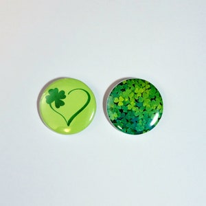May include: Two round green buttons with a shamrock design. One button features a shamrock inside a heart shape, while the other button is covered in shamrocks.