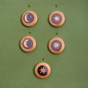 May include: Five round wooden buttons with celestial designs. Each button features a different design: a crescent moon and star, a sun with rays, a face on a crescent moon, a sun with rays, and a star. The designs are in white and blue.