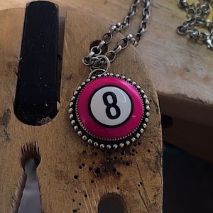 May include: A silver chain necklace with a pendant featuring a pink and white eight ball design. The pendant has a silver beaded border and a black number eight. The necklace is displayed on a wooden surface.